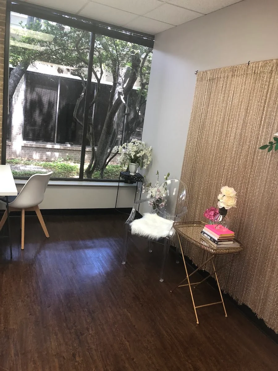 Business Lounge Dallas boutique private office with dark hardwood floors, white Eames-style desk chair, ghost chair with faux-fur cushion, gold side table with pink books and flowers, and sparkly gold curtain divider by a garden-view window