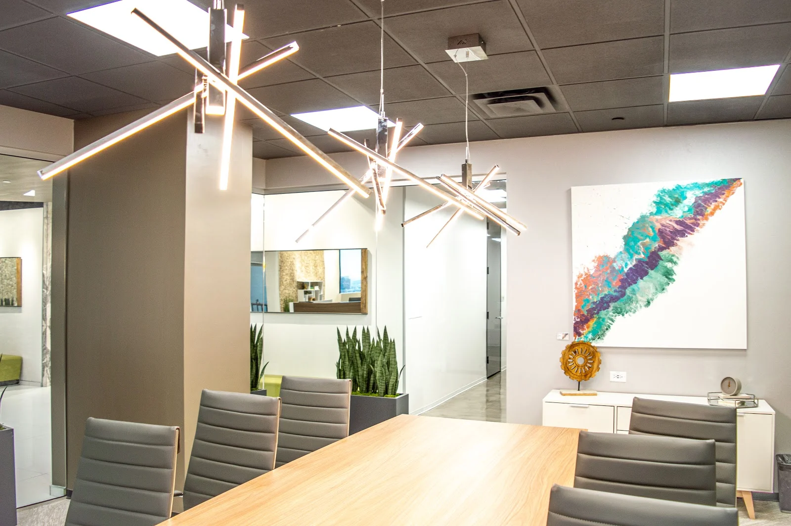Lucid Private Offices Uptown Dallas modern conference room with light oak table, grey leather executive chairs, dramatic angular chrome LED light fixtures, colorful teal-purple-orange abstract art, snake plants in planter, and glass partition