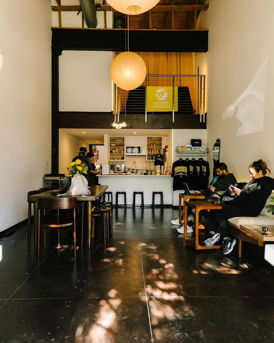 Hyphen Space double-height converted industrial space with exposed timber rafters, large paper lantern pendant, Hyphen Studios yellow banner on mezzanine, open kitchen-coffee bar with black stools, members working at dark wood tables, and natural light patterns on polished concrete floor