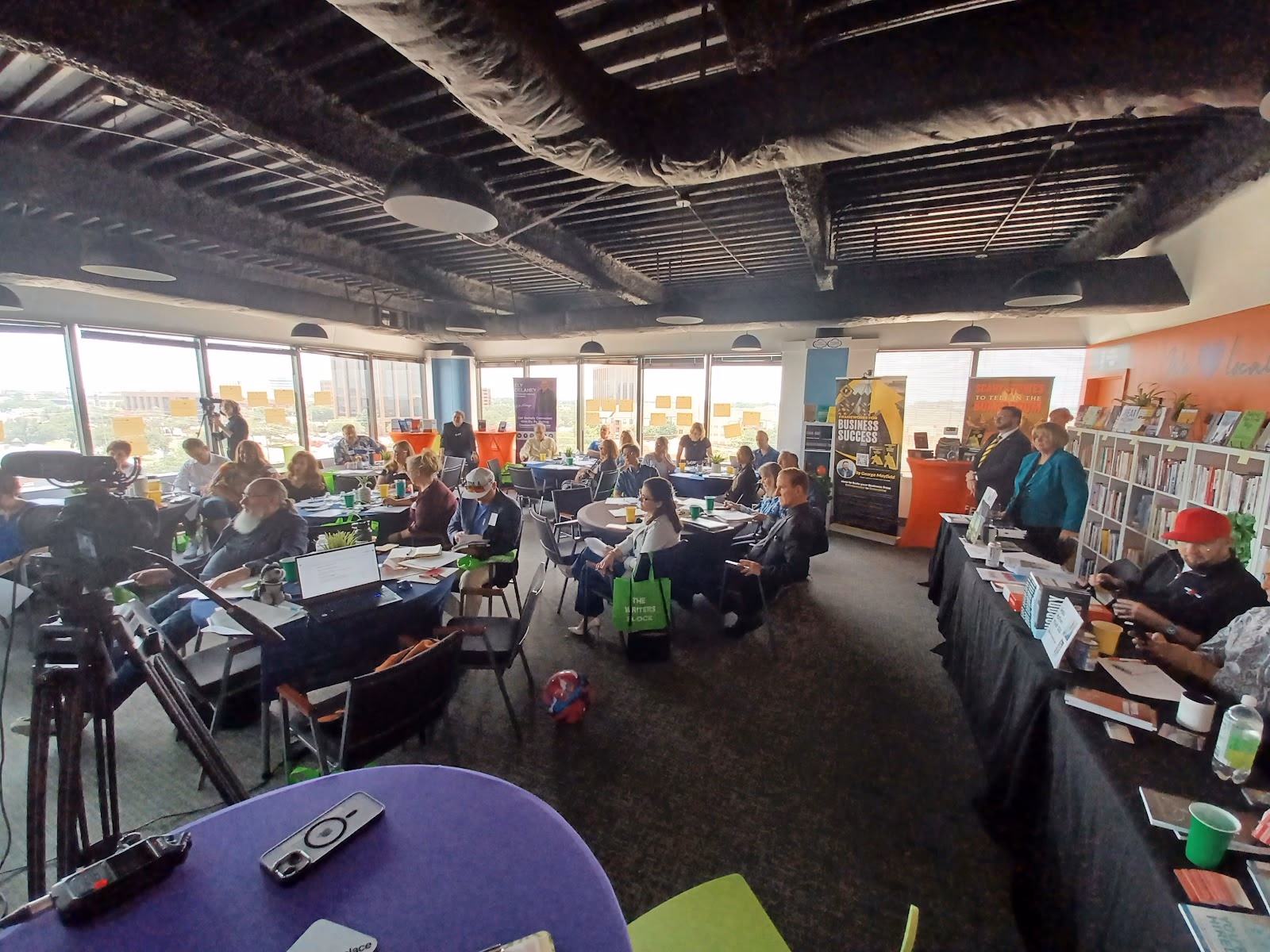 The Meeting Place packed high-floor event space with panoramic windows, dozens of attendees at round and rectangular tables during a workshop presentation, orange accent wall with bookshelf, exposed black ceiling with speakers, and professional video equipment