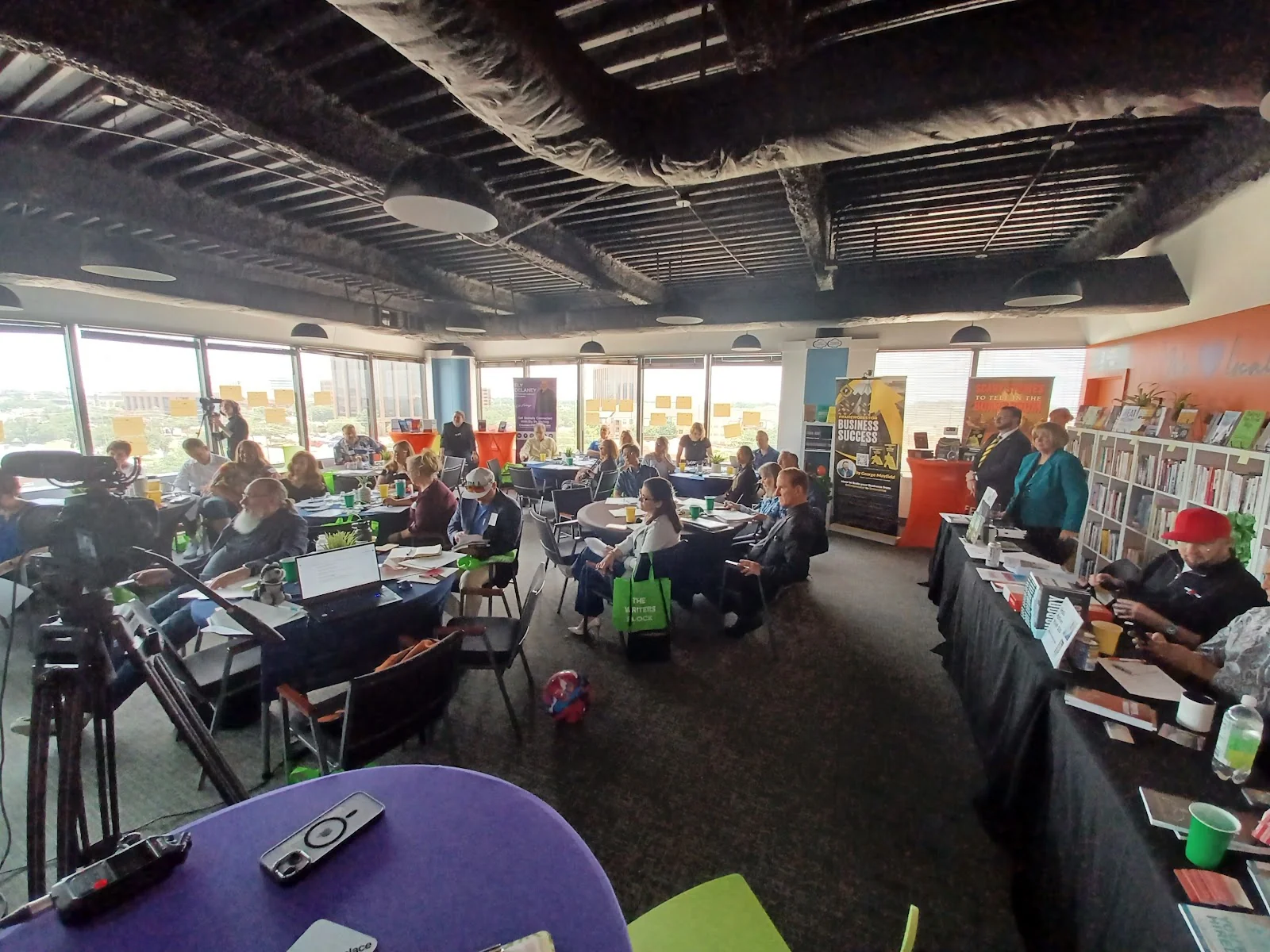 The Meeting Place packed high-floor event space with panoramic windows, dozens of attendees at round and rectangular tables during a workshop presentation, orange accent wall with bookshelf, exposed black ceiling with speakers, and professional video equipment