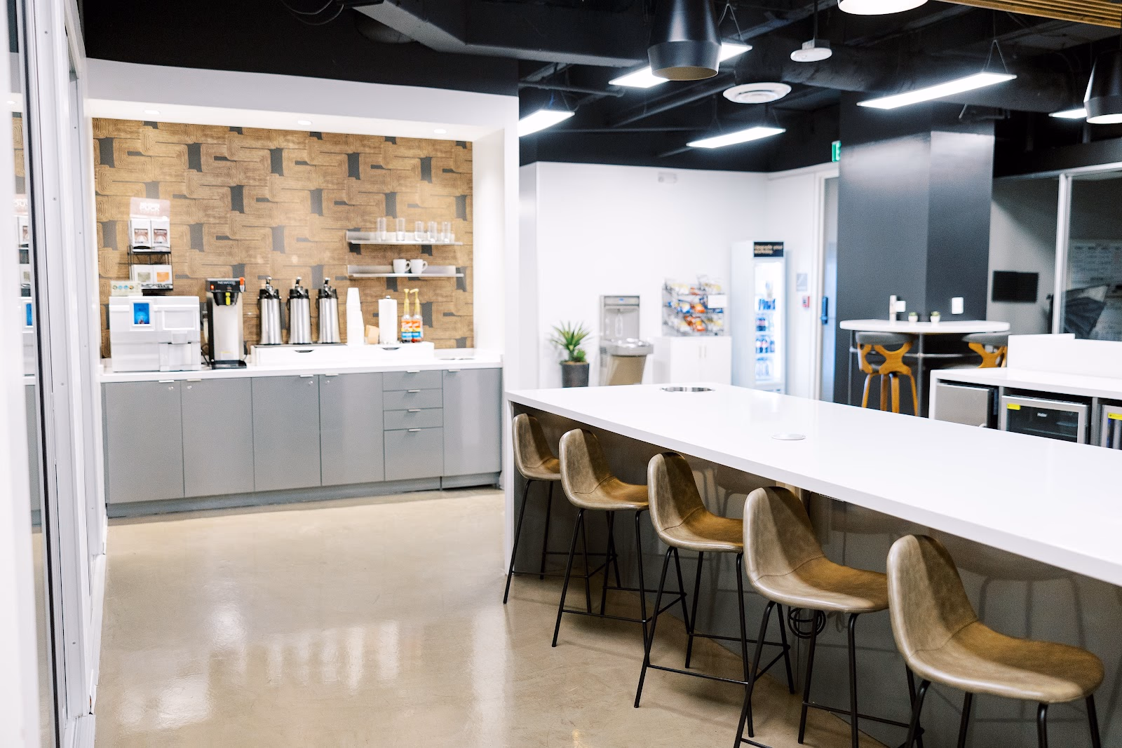 Lucid Private Offices Cole Avenue kitchen and break area with geometric wood-and-grey wallpaper accent wall, stainless coffee urns on white counter, grey cabinets, tan leather bar stools along a white communal island, and black ceiling with pendant lights