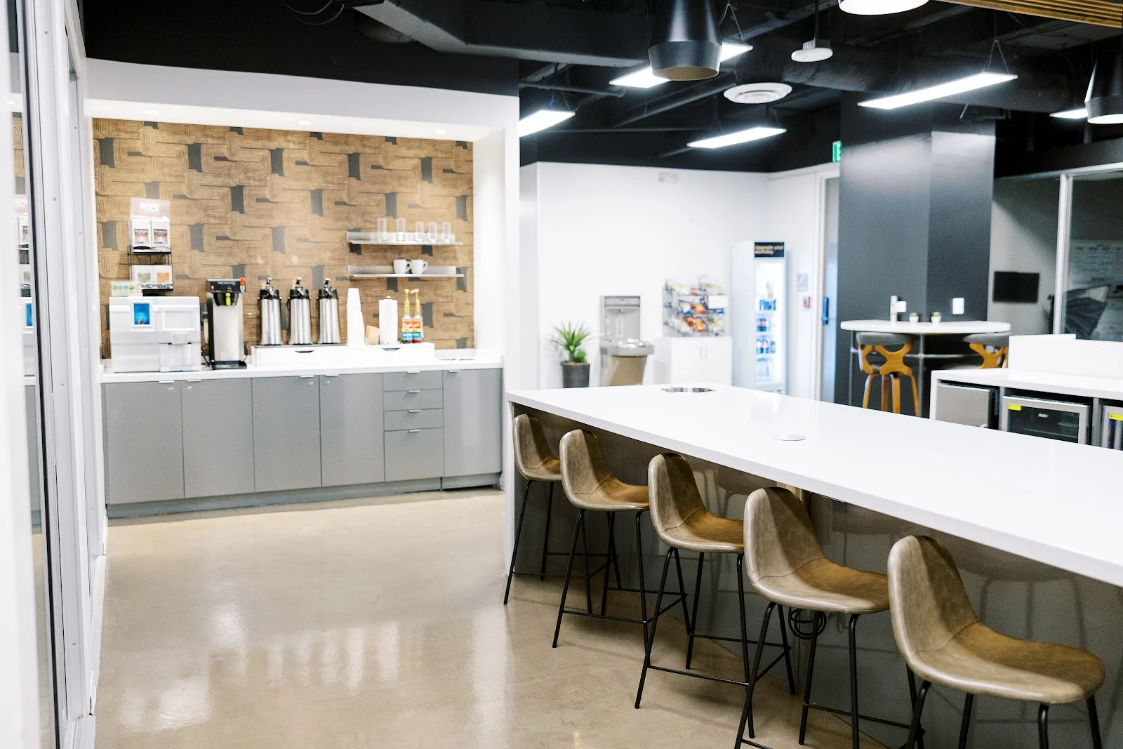 Lucid Private Offices Cole Avenue kitchen and break area with geometric wood-and-grey wallpaper accent wall, stainless coffee urns on white counter, grey cabinets, tan leather bar stools along a white communal island, and black ceiling with pendant lights