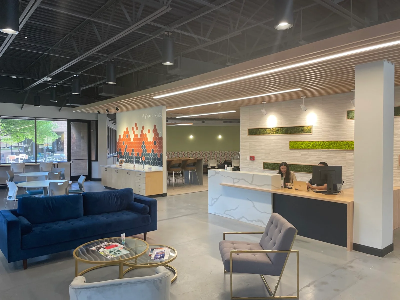 HEXA Coworking lobby with navy velvet sofa, gold nesting coffee tables, grey tufted accent chair, white marble-front reception desk with wood slat ceiling above, orange hexagonal tile mural, moss wall panels, and polished concrete floors
