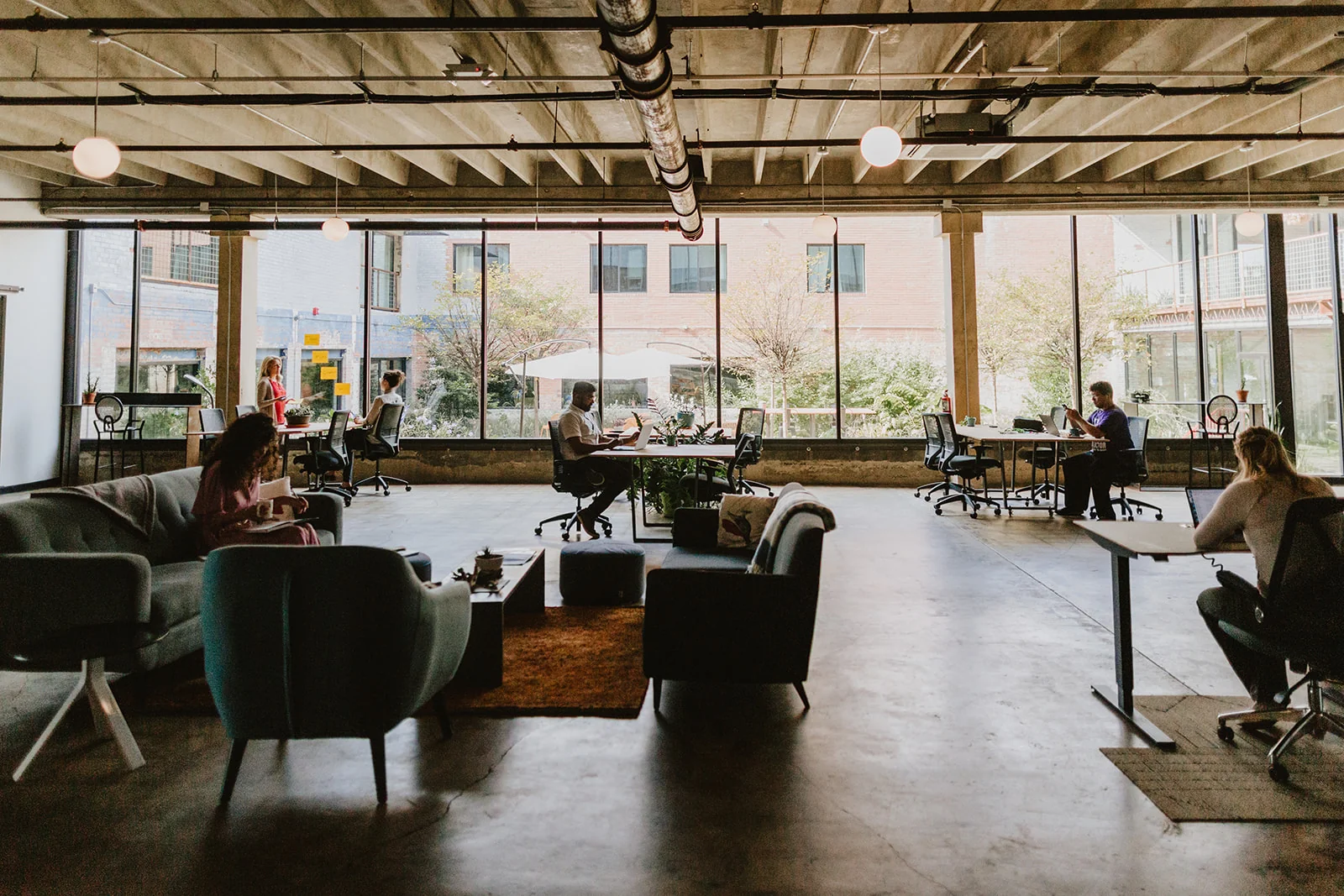 Good Coworking spacious industrial interior with floor-to-ceiling windows overlooking trees and brick buildings, polished concrete floors, grey armchairs and sofas on a warm area rug, members at ergonomic desk stations, exposed ceiling beams, and globe pendant lights