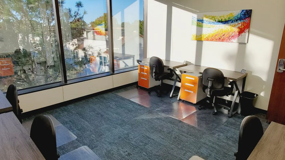 TRUE Space Dallas Uptown private office with two L-shaped desks, ergonomic black chairs, orange mobile filing cabinets, colorful abstract wave painting on the wall, floor-to-ceiling window with tree-lined view, and blue-green textured carpet