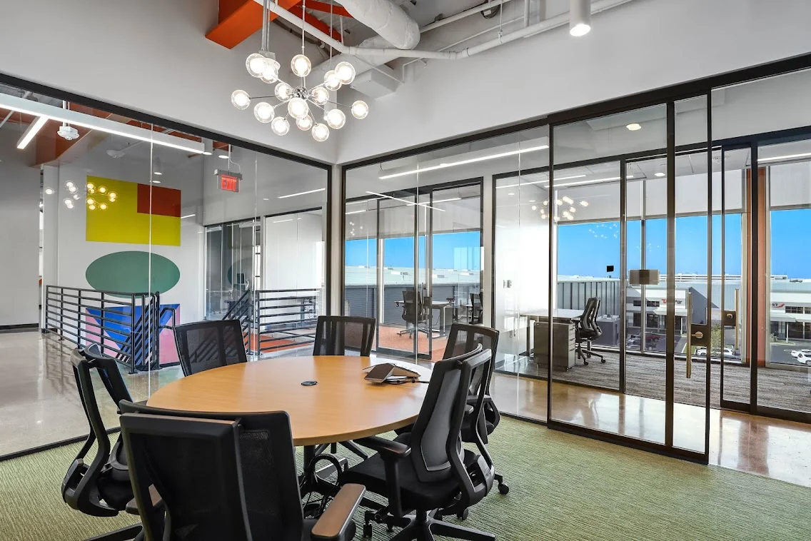 Venture X Braniff Centre conference room with round wood table and black mesh chairs, sputnik globe chandelier, bold yellow-red-teal geometric art on white wall, glass-walled private offices with skyline view, green carpet, and exposed ceiling ductwork with orange accents