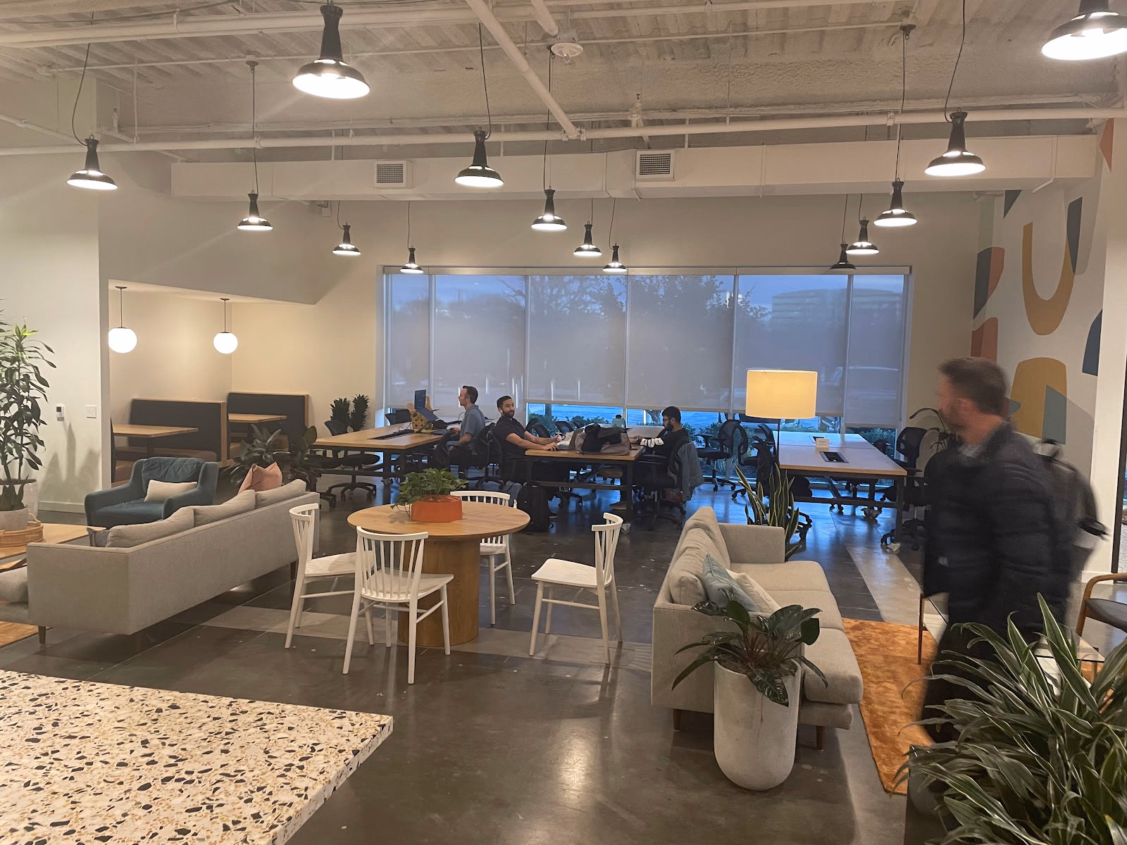 Common Desk Richardson open coworking lounge with industrial pendant lights, grey sofas and armchairs, white Windsor chairs at round wood table, terrazzo kitchen counter, colorful abstract mural on far wall, potted plants, and members working at communal tables by large shaded windows