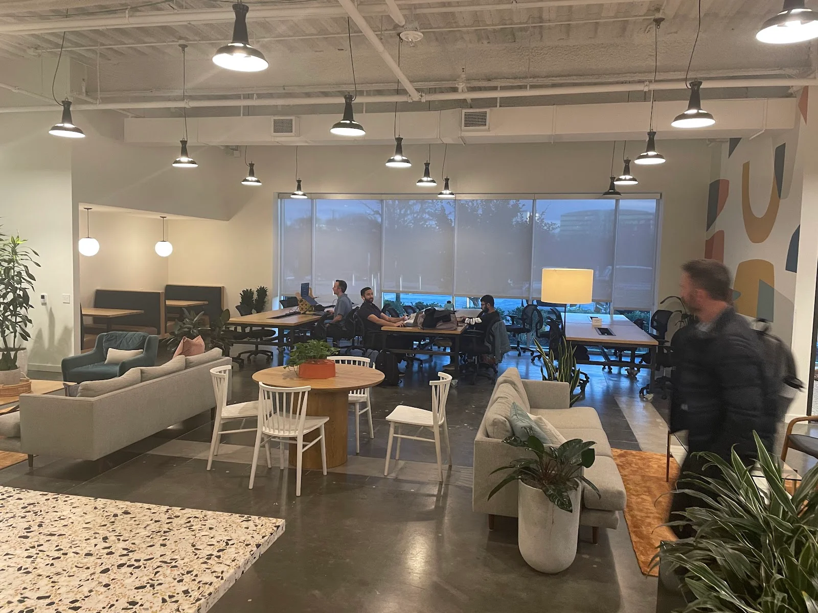 Common Desk Richardson open coworking lounge with industrial pendant lights, grey sofas and armchairs, white Windsor chairs at round wood table, terrazzo kitchen counter, colorful abstract mural on far wall, potted plants, and members working at communal tables by large shaded windows