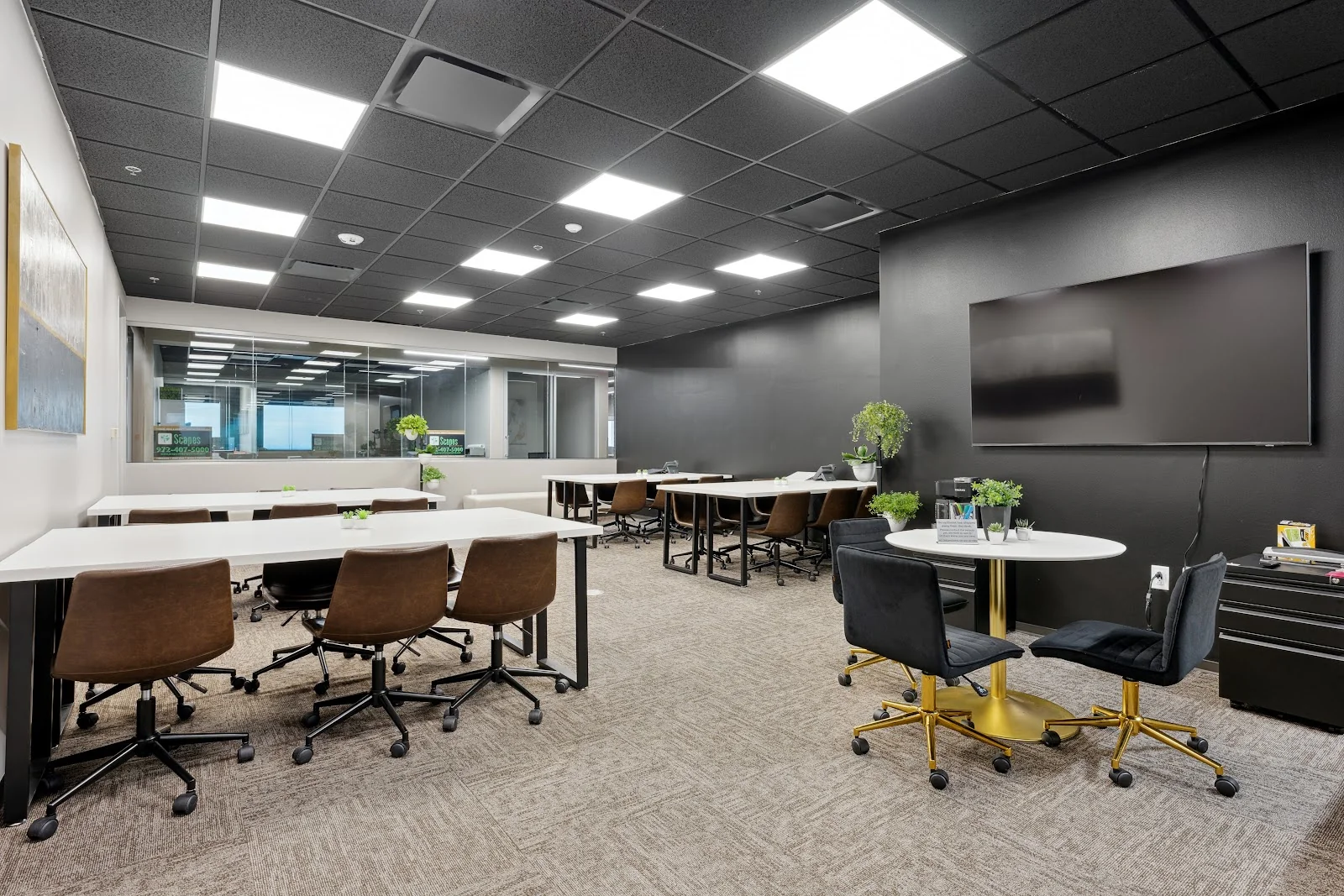 Lucid Private Offices Northpark coworking room with dark ceiling panels, bright LED lighting, white communal tables with brown leather rolling chairs, dark accent wall with large flat-screen TV, small round table with navy velvet chairs on gold bases, and potted green plants