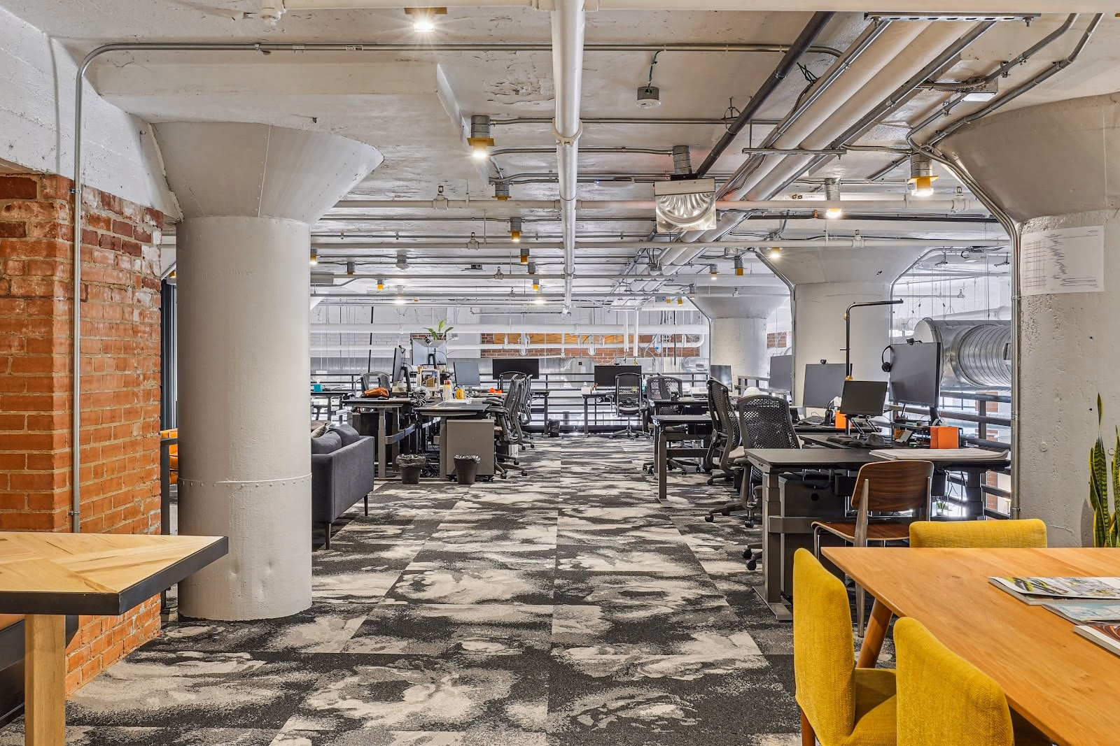 Common Desk West End deep industrial workspace with exposed brick columns, concrete pillars, patterned grey carpet, rows of dedicated desks with dual monitors and ergonomic mesh chairs, yellow accent chairs at a wood communal table, and exposed ceiling pipes
