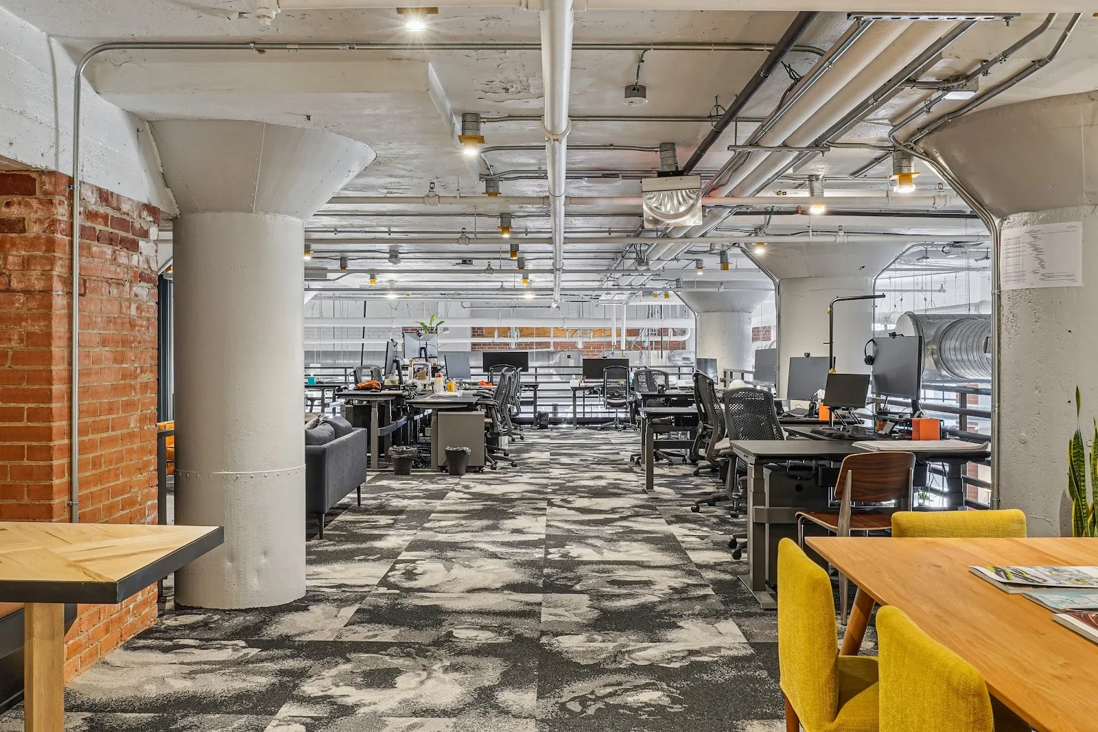 Common Desk West End deep industrial workspace with exposed brick columns, concrete pillars, patterned grey carpet, rows of dedicated desks with dual monitors and ergonomic mesh chairs, yellow accent chairs at a wood communal table, and exposed ceiling pipes