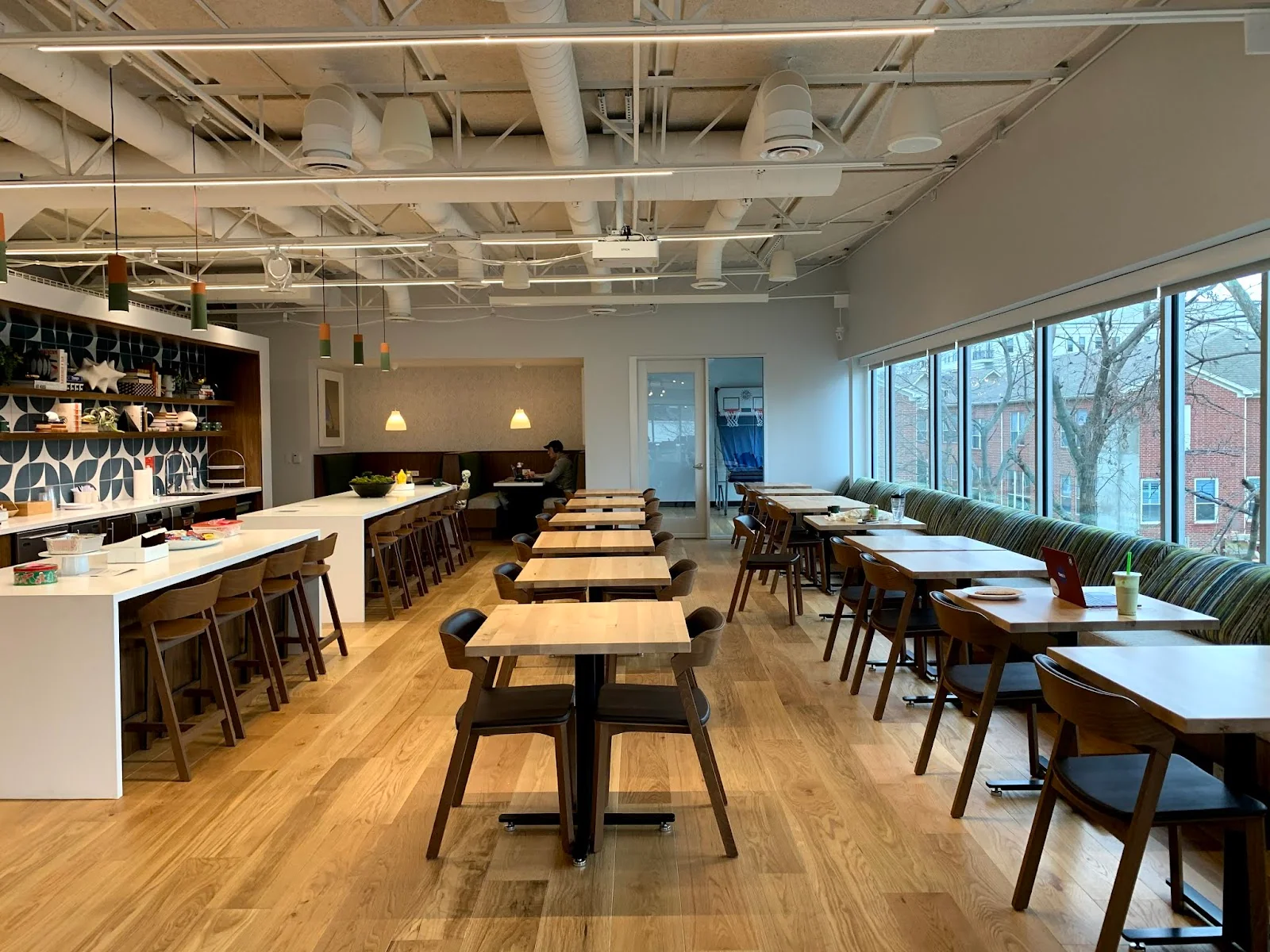 Work214 bright cafe-style workspace with light oak hardwood floors, rows of butcher-block tables with dark wood chairs, white kitchen island with bar stools, blue geometric tile backsplash with open shelving, colorful pendant lights, green striped banquette along floor-to-ceiling windows, and exposed white ductwork ceiling