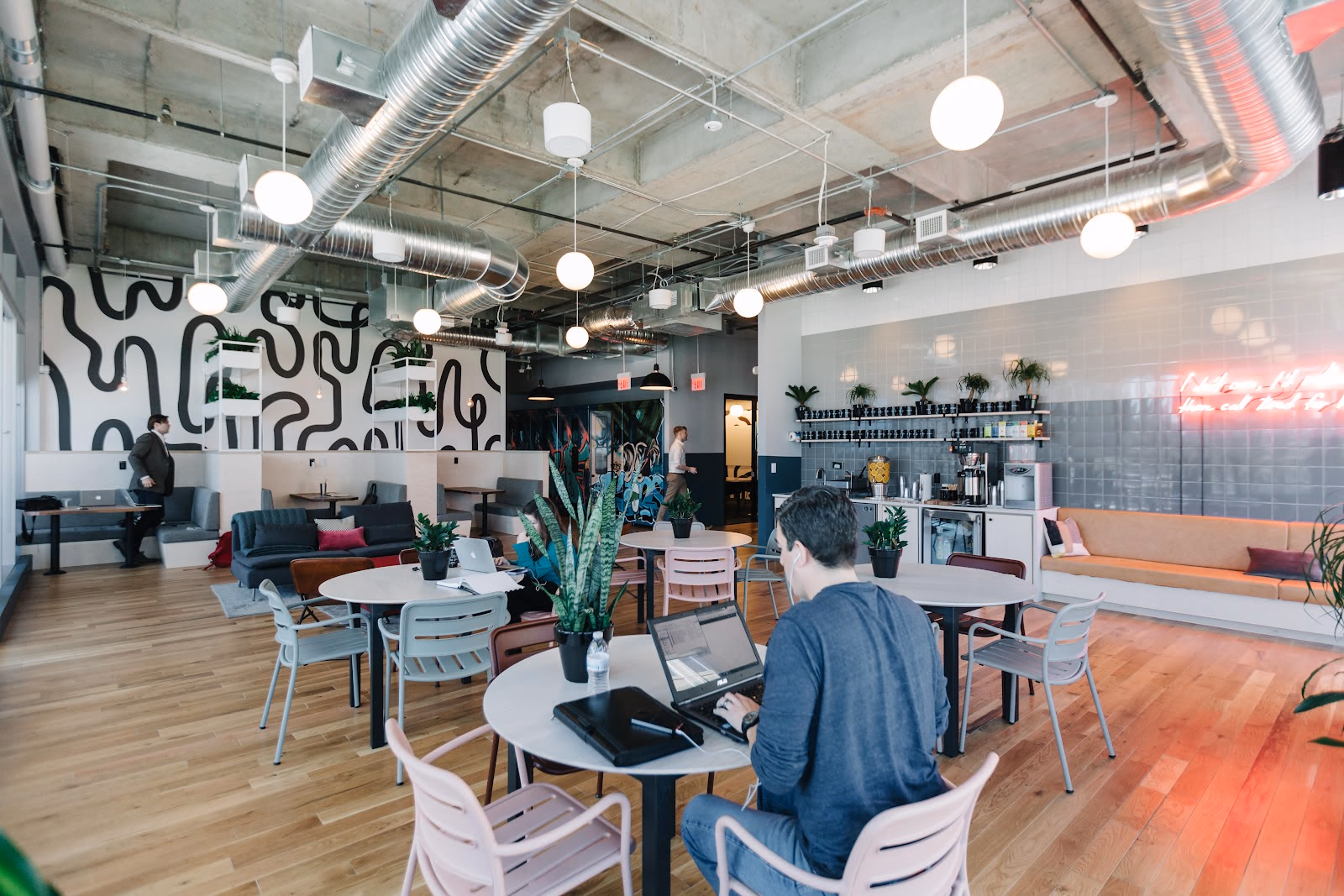 WeWork McKinney Avenue open lounge with bold black-and-white abstract mural, exposed ductwork and globe pendant lights, round cafe tables with pastel chairs, members working on laptops, grey booth seating with colorful pillows, coffee bar with neon sign, hardwood floors, and potted plants