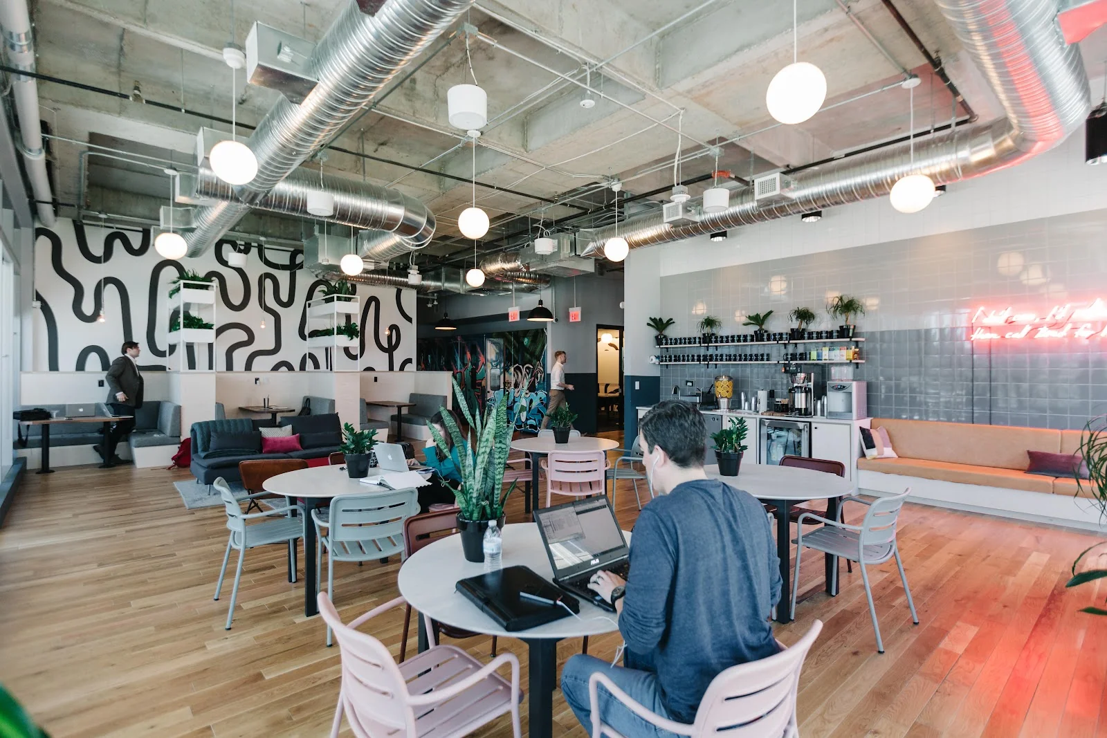 WeWork McKinney Avenue open lounge with bold black-and-white abstract mural, exposed ductwork and globe pendant lights, round cafe tables with pastel chairs, members working on laptops, grey booth seating with colorful pillows, coffee bar with neon sign, hardwood floors, and potted plants
