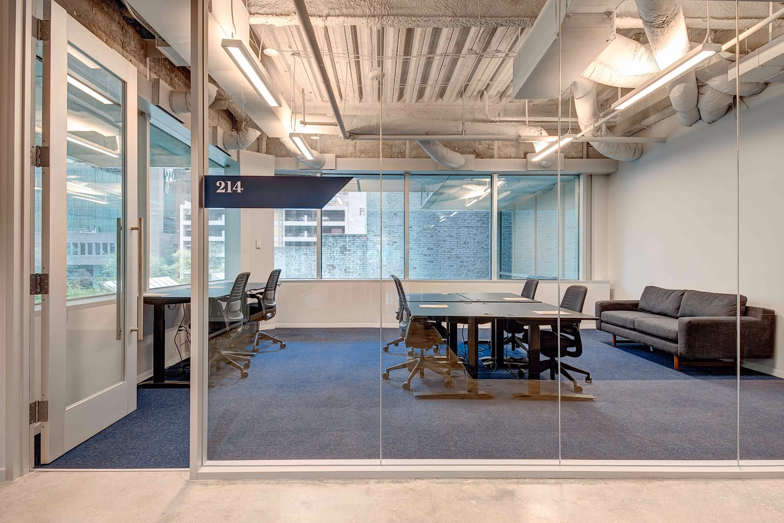 The Cue Arts District glass-walled conference room labeled 214 with blue carpet, dark conference table surrounded by ergonomic mesh chairs, grey sofa along the wall, exposed concrete ceiling with linear LED fixtures and wrapped ductwork, and city-view windows