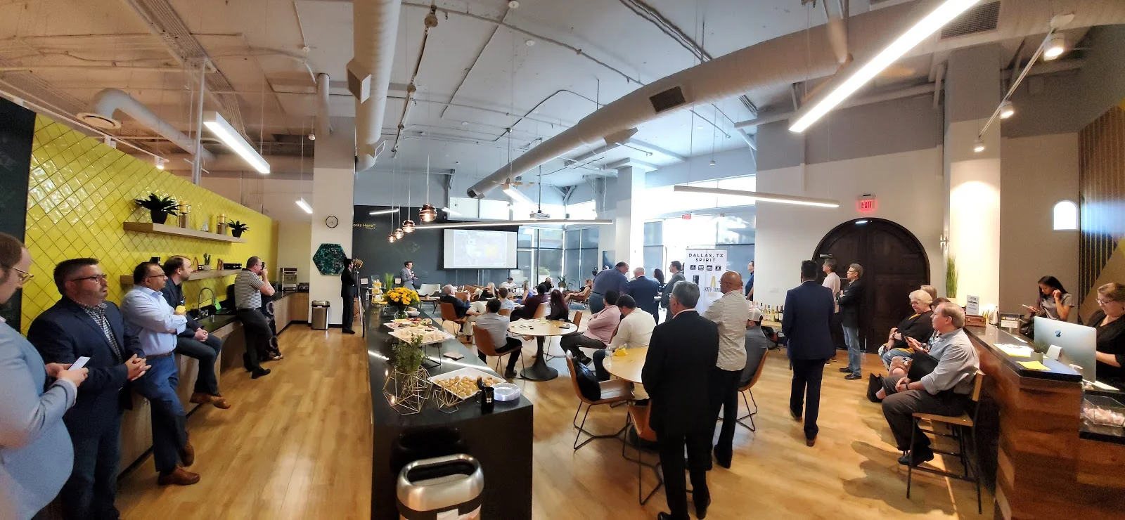 Venture X Galleria networking event in large open space with bright yellow tile kitchen wall, copper pendant lights, presentation screen, round tables with dozens of professionals mingling, arched doorway, warm hardwood floors, and exposed white ductwork ceiling