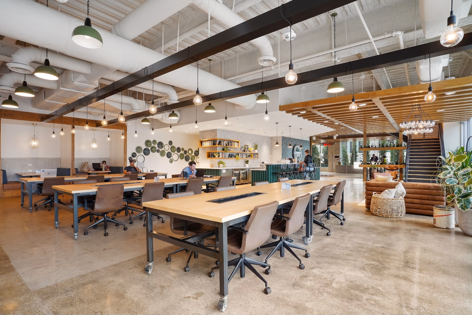 Common Desk Energy Square warm industrial coworking hall with long butcher-block communal tables on steel frames, brown leather rolling chairs, green dome pendant lights and Edison bulbs hanging from black steel beams, decorative ceramic plates on wall, green-tiled coffee bar, wooden slat ceiling, and polished concrete floors
