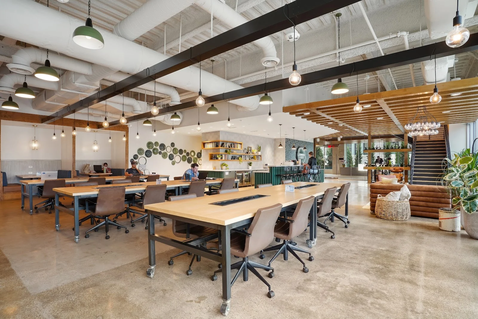 Common Desk Energy Square warm industrial coworking hall with long butcher-block communal tables on steel frames, brown leather rolling chairs, green dome pendant lights and Edison bulbs hanging from black steel beams, decorative ceramic plates on wall, green-tiled coffee bar, wooden slat ceiling, and polished concrete floors