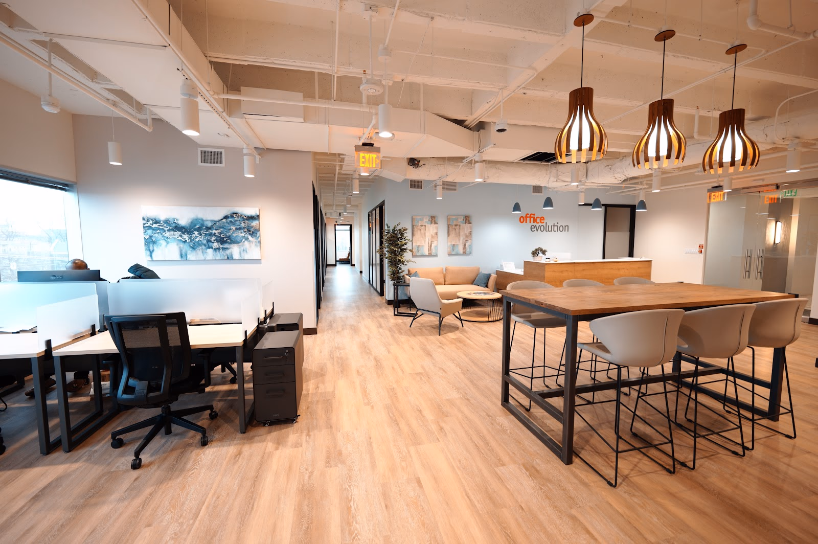 Office Evolution Turtle Creek modern open-plan office with light blonde wood-look vinyl floors, hot-desk stations with privacy screens, high communal table with cream bucket chairs, sculptural wooden gourd pendant lights, blue abstract ocean art on light blue walls, and exposed white ceiling ductwork