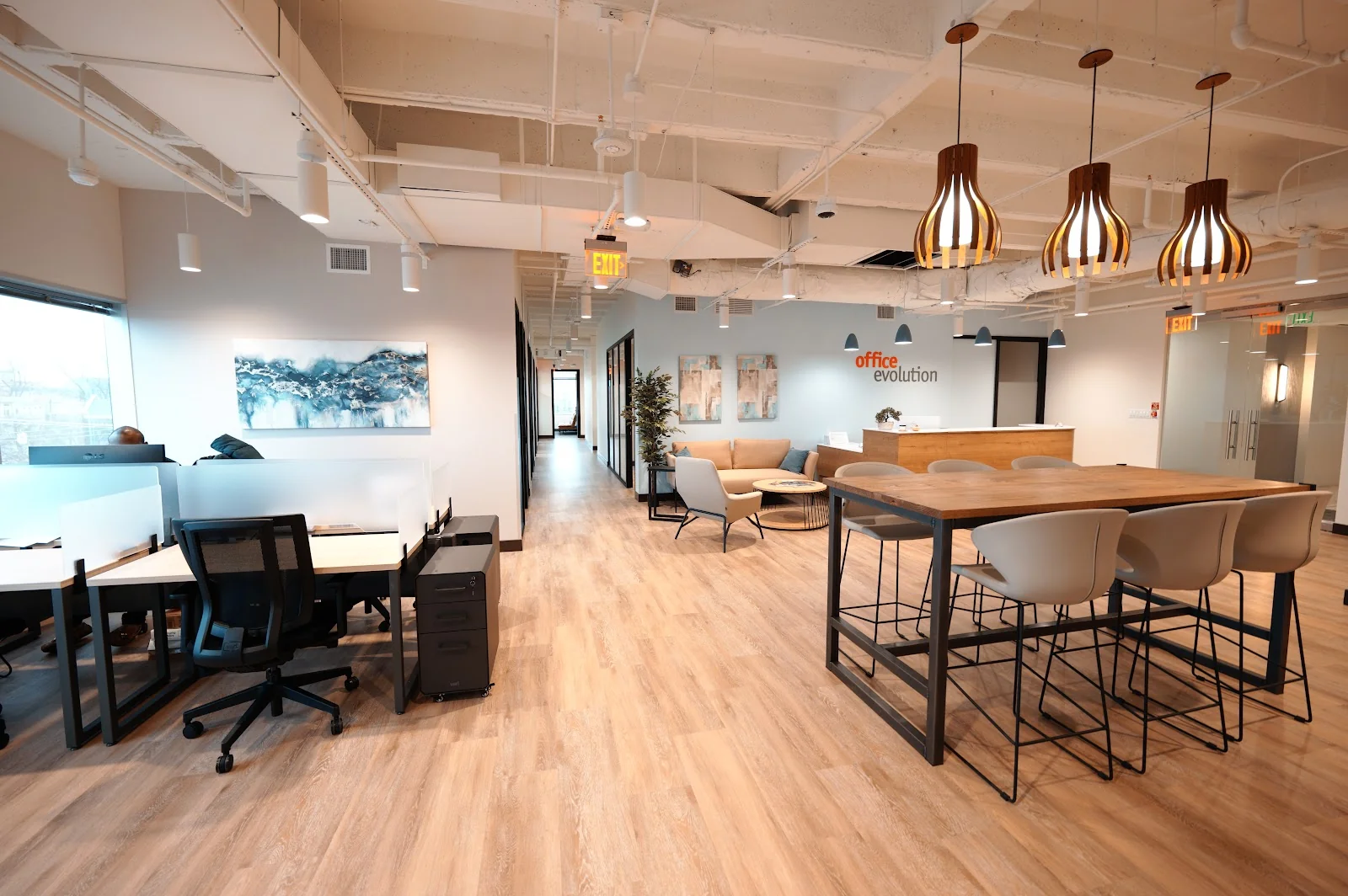 Office Evolution Turtle Creek modern open-plan office with light blonde wood-look vinyl floors, hot-desk stations with privacy screens, high communal table with cream bucket chairs, sculptural wooden gourd pendant lights, blue abstract ocean art on light blue walls, and exposed white ceiling ductwork