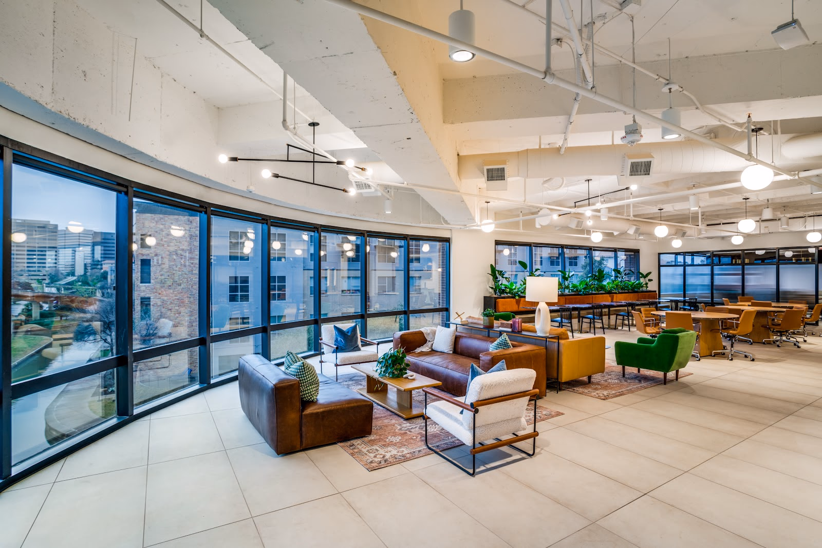 Foster Coworking Canal Centre luxurious curved-glass lounge with floor-to-ceiling black-framed windows overlooking a canal at dusk, brown leather sofas and cream armchairs on vintage Persian rug, green wingback chair, exposed white ceiling with industrial ductwork, globe lights, and plant dividers