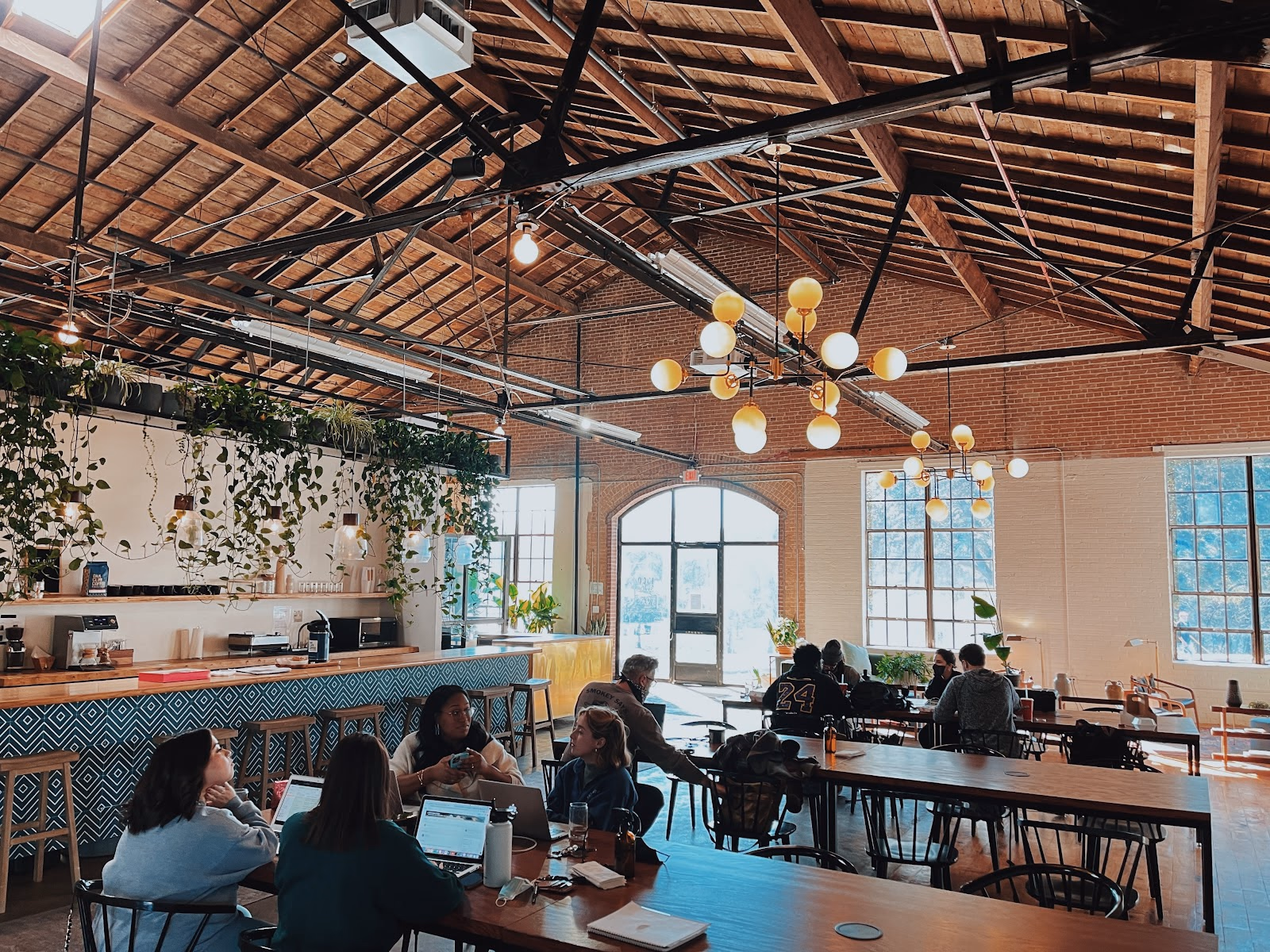 Wax Space converted warehouse with soaring timber-truss ceiling, exposed brick walls, large arched windows flooding natural light, hanging pothos plants along a teal diamond-pattern tiled bar counter, globe pendant chandeliers, and members working at wooden communal tables
