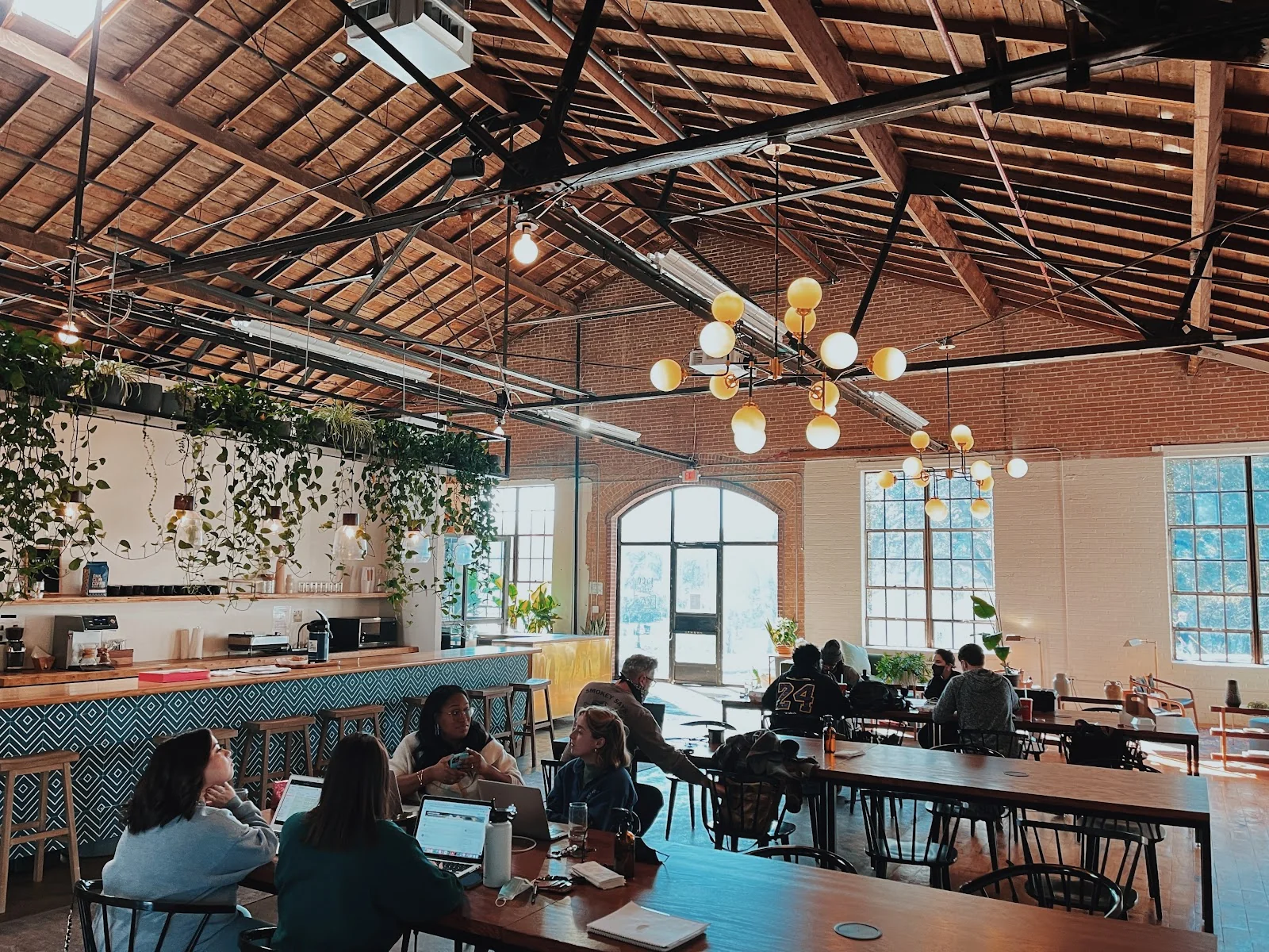Wax Space converted warehouse with soaring timber-truss ceiling, exposed brick walls, large arched windows flooding natural light, hanging pothos plants along a teal diamond-pattern tiled bar counter, globe pendant chandeliers, and members working at wooden communal tables