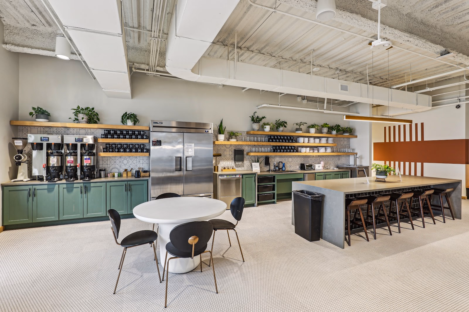 Common Desk Dallas Arts District communal kitchen with green-painted lower cabinets, multiple coffee brewers on wooden shelves with potted plants, stainless steel refrigerator, geometric tile backsplash, white tulip table with black chairs, concrete island bar with wood stools, and orange-brown striped accent wall