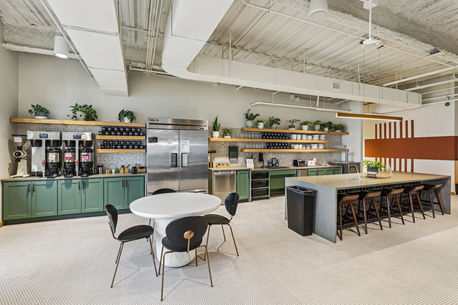 Common Desk Dallas Arts District communal kitchen with green-painted lower cabinets, multiple coffee brewers on wooden shelves with potted plants, stainless steel refrigerator, geometric tile backsplash, white tulip table with black chairs, concrete island bar with wood stools, and orange-brown striped accent wall