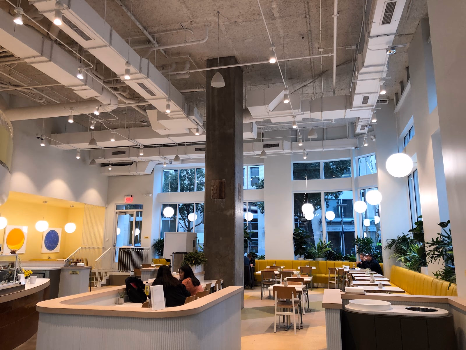 Hatchways Workspace bright cafe-style interior with soaring double-height ceilings, exposed concrete column, globe pendant lights, yellow banquette seating, white cafe tables and chairs, yellow accent wall with circular artwork, and large windows showing Victory Park street views