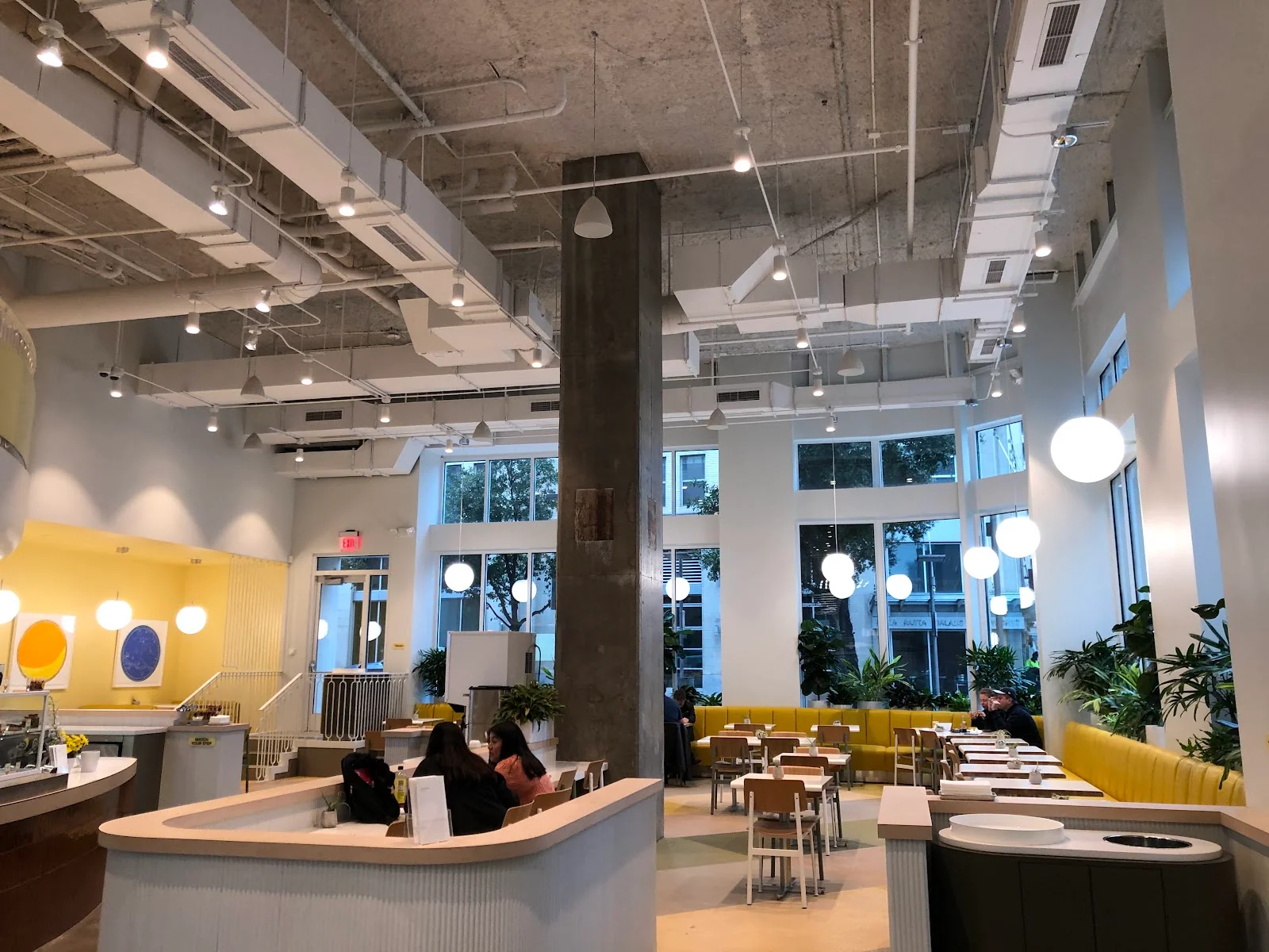 Hatchways Workspace bright cafe-style interior with soaring double-height ceilings, exposed concrete column, globe pendant lights, yellow banquette seating, white cafe tables and chairs, yellow accent wall with circular artwork, and large windows showing Victory Park street views