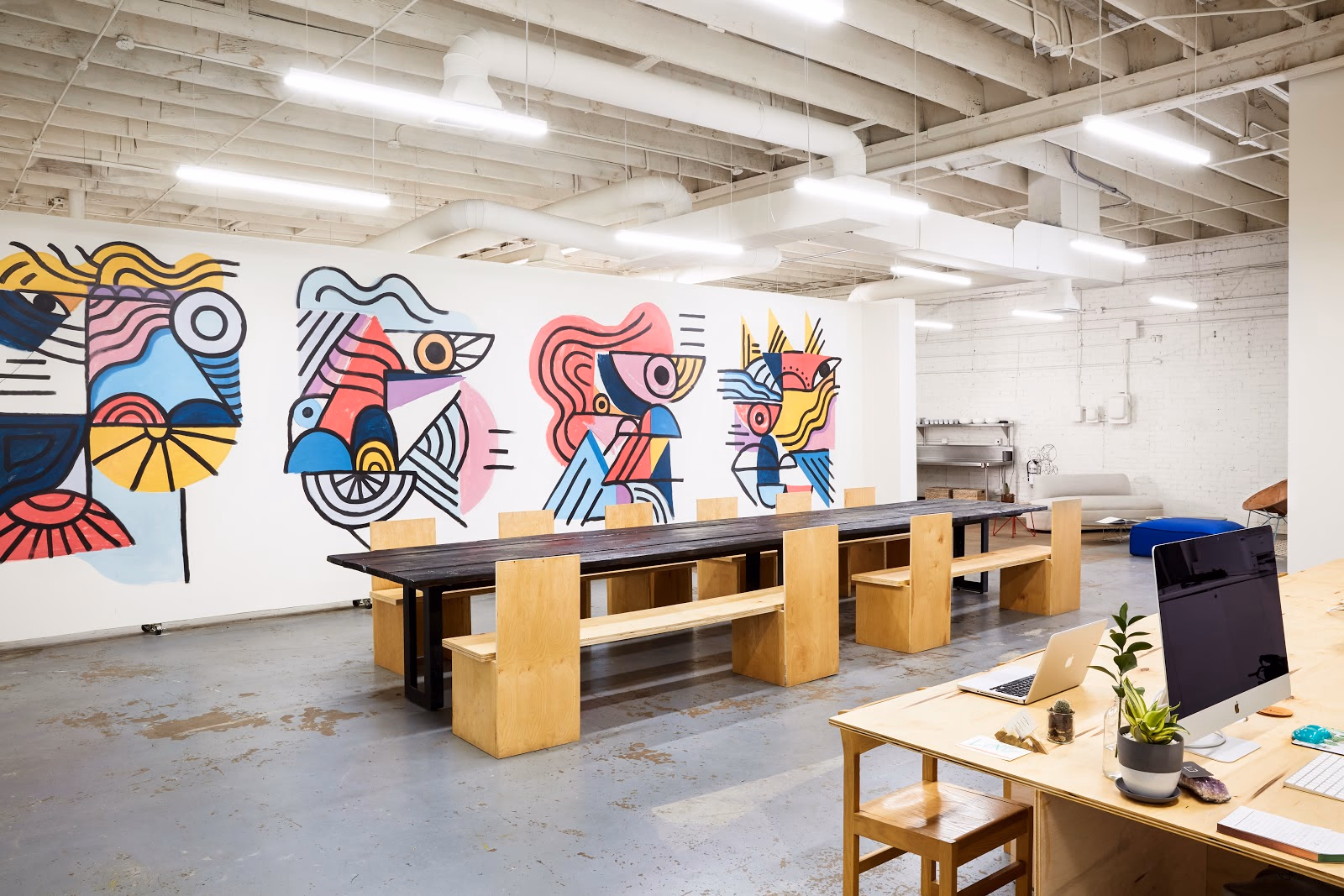FLOCC creative studio with polished concrete floors, white-painted exposed ceiling beams, long dark reclaimed-wood communal table with plywood box-style stools, massive colorful abstract mural spanning the entire wall in bold pinks blues yellows and blacks, and natural wood desk with laptop and plants