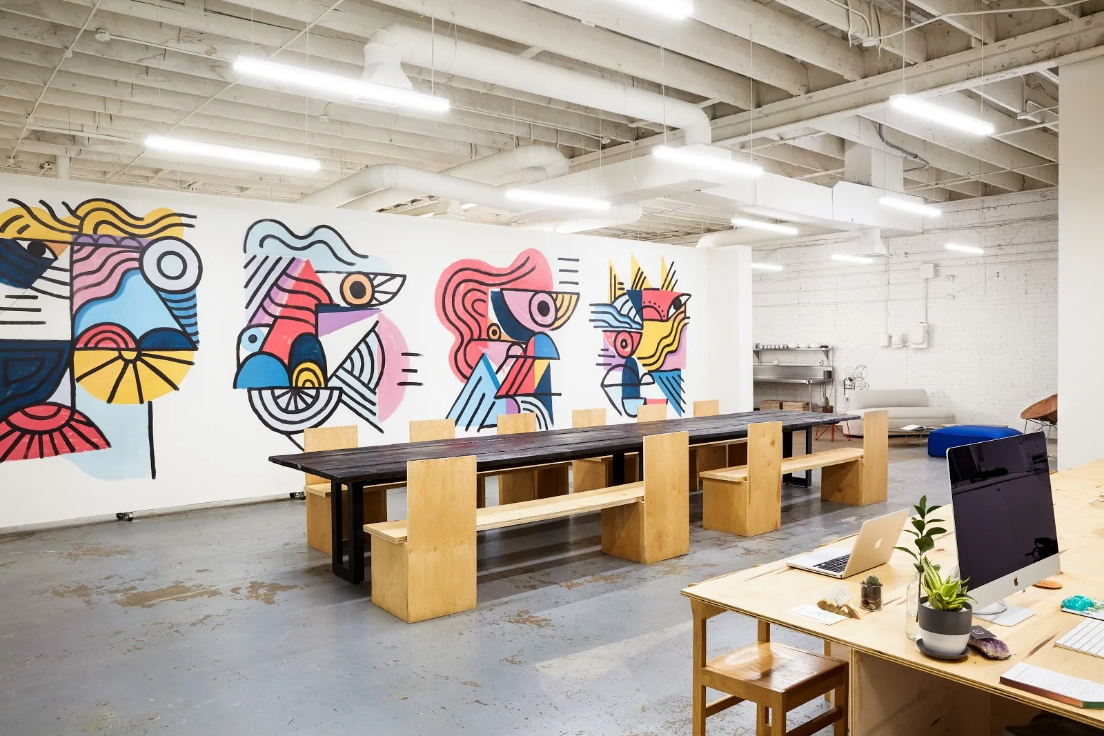 FLOCC creative studio with polished concrete floors, white-painted exposed ceiling beams, long dark reclaimed-wood communal table with plywood box-style stools, massive colorful abstract mural spanning the entire wall in bold pinks blues yellows and blacks, and natural wood desk with laptop and plants