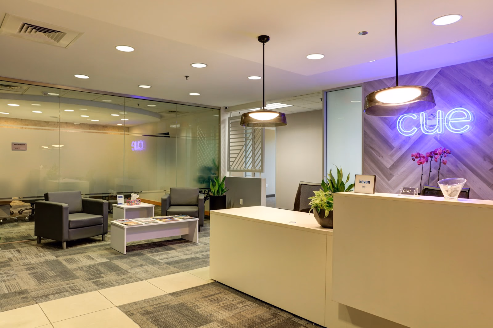 The Cue North Dallas polished reception area with white desk, herringbone grey wood accent wall featuring glowing blue neon cue sign, grey leather armchairs in waiting area, orchid and potted plants on desk, glass-walled conference room visible behind, and grey patterned carpet