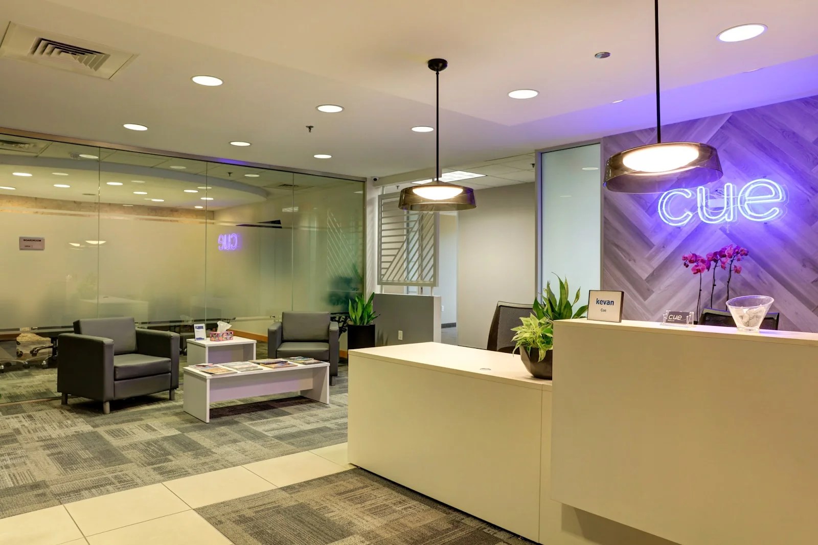 The Cue North Dallas polished reception area with white desk, herringbone grey wood accent wall featuring glowing blue neon cue sign, grey leather armchairs in waiting area, orchid and potted plants on desk, glass-walled conference room visible behind, and grey patterned carpet