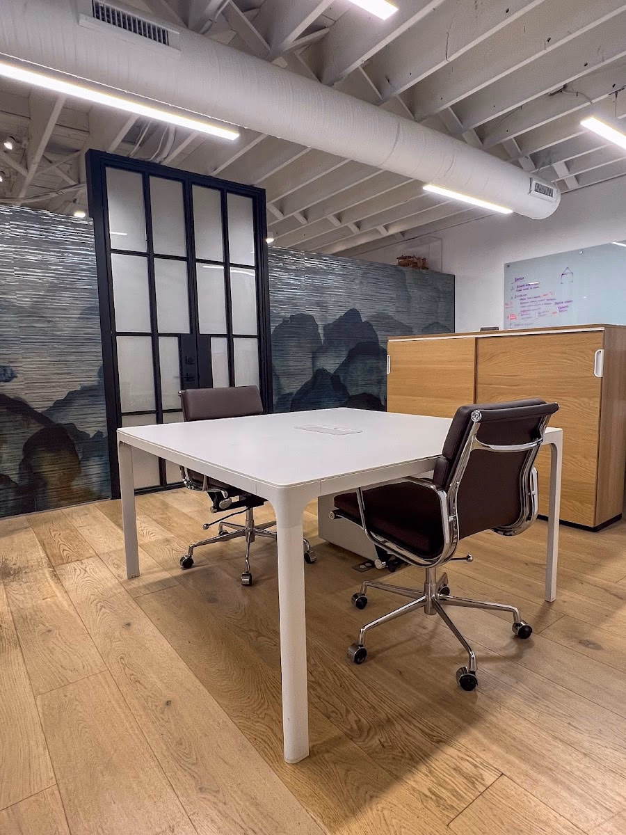 Spryrocket Station private office with warm oak hardwood floors, white desk with two brown leather Eames-style chairs, dramatic blue-grey mountain landscape wallpaper mural, black-framed frosted glass partition door, oak storage cabinet, whiteboard, and exposed white ceiling with linear LED fixtures