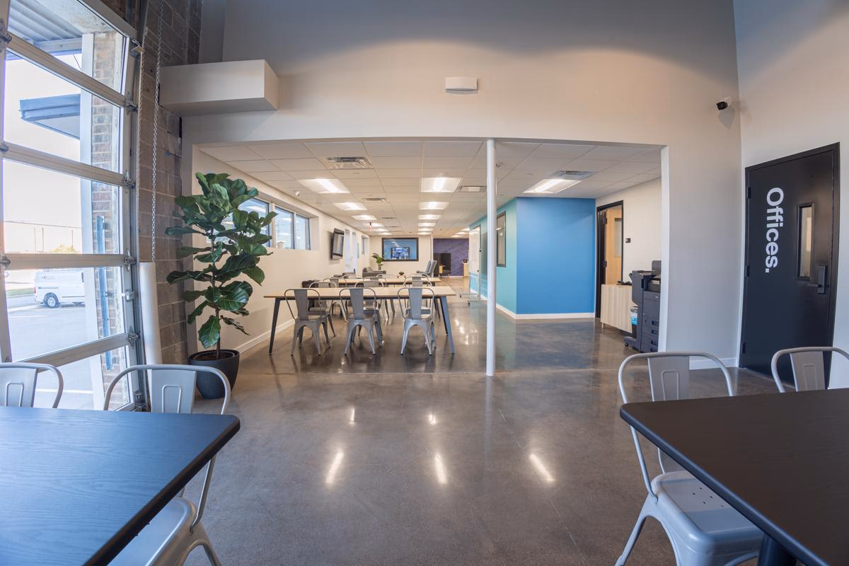 Polygon converted industrial space with roll-up garage door windows flooding natural light, polished concrete floors, silver Tolix-style metal chairs at dark communal tables, fiddle-leaf fig plant, teal accent wall, Offices sign on dark door, and clerestory windows