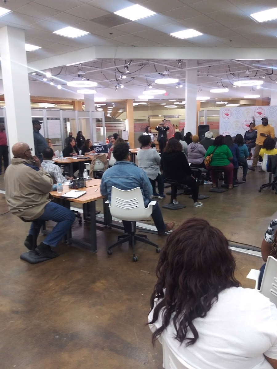 The Dallas Entrepreneur Center at RedBird community workshop with fluorescent ring lights, wood-framed glass partitions, polished concrete floors, rows of attendees seated at tables watching a presentation, and orange-coral accent branding banners on walls
