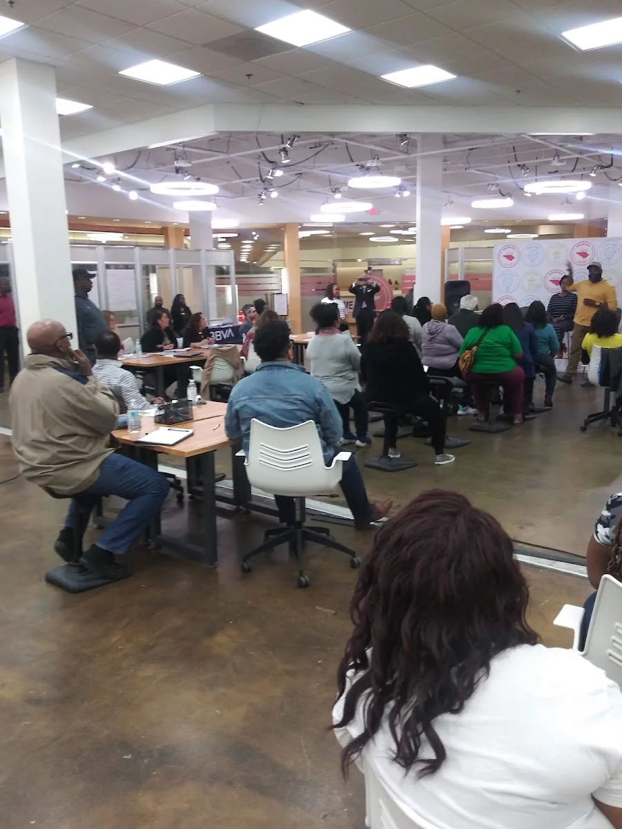 The Dallas Entrepreneur Center at RedBird community workshop with fluorescent ring lights, wood-framed glass partitions, polished concrete floors, rows of attendees seated at tables watching a presentation, and orange-coral accent branding banners on walls
