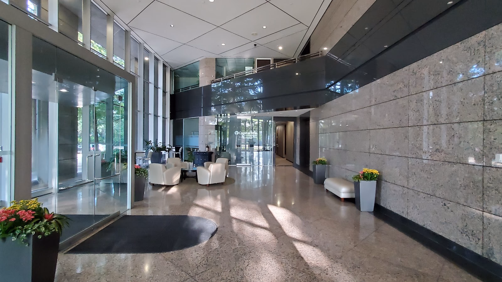 TRUE Space Dallas Far North grand corporate lobby with double-height glass curtain wall, polished granite floors and stone accent walls, white club chairs arranged around a small table, recessed lighting, and potted flowers at the entrance