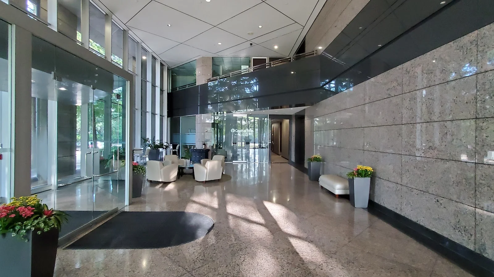 TRUE Space Dallas Far North grand corporate lobby with double-height glass curtain wall, polished granite floors and stone accent walls, white club chairs arranged around a small table, recessed lighting, and potted flowers at the entrance