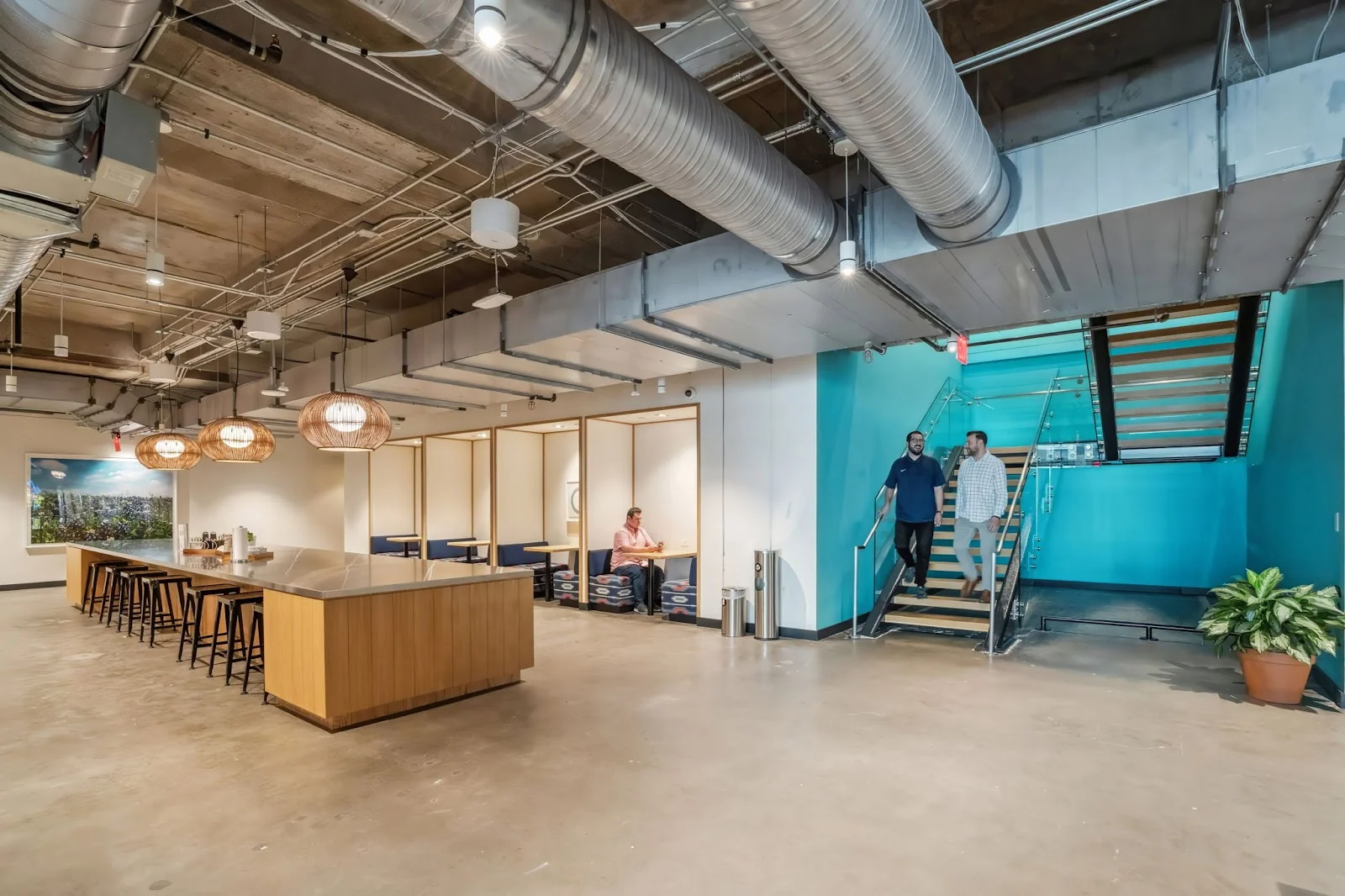 Workbox Dallas Victory Park industrial lobby with exposed ductwork, wood-paneled kitchen island with black bar stools, woven rattan pendant lights, semi-private booth seating along wall, teal accent staircase with two men descending, and polished concrete floors