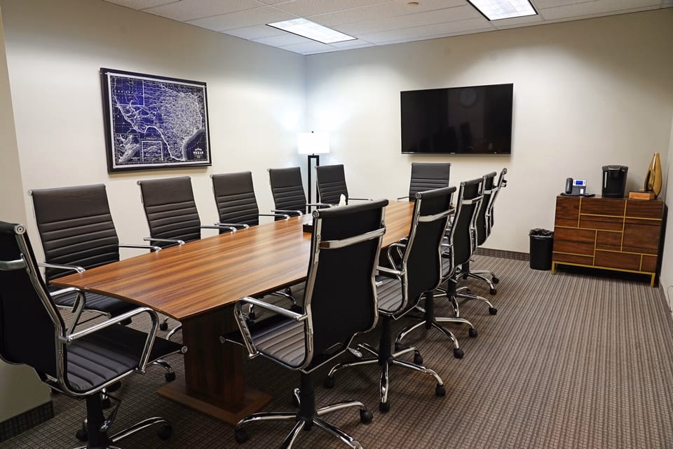 ExecutiveWorkspace Uptown Dallas traditional boardroom with long walnut conference table, black leather high-back executive chairs, framed vintage Texas map on wall, wall-mounted flat-screen TV, walnut credenza with Keurig, and striped commercial carpet