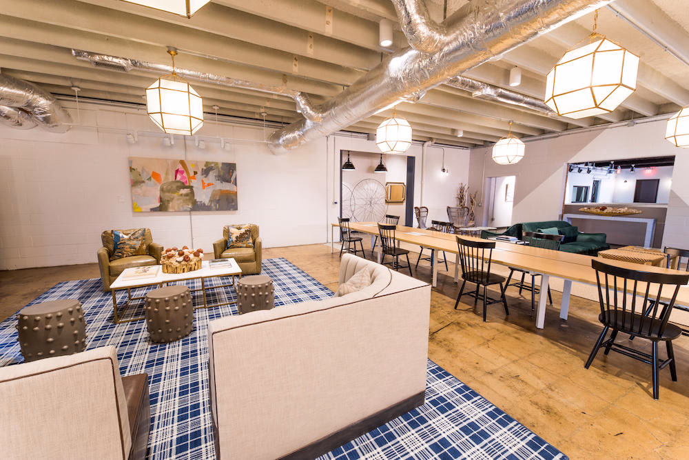 The Slate industrial-chic lounge with exposed ceiling ducts and beams, geometric pendant lights, cream sofas and olive wingback chairs on blue plaid rug, long wooden communal table with black Windsor chairs, polished concrete floors, and abstract art on white brick wall
