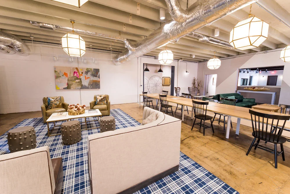 The Slate industrial-chic lounge with exposed ceiling ducts and beams, geometric pendant lights, cream sofas and olive wingback chairs on blue plaid rug, long wooden communal table with black Windsor chairs, polished concrete floors, and abstract art on white brick wall