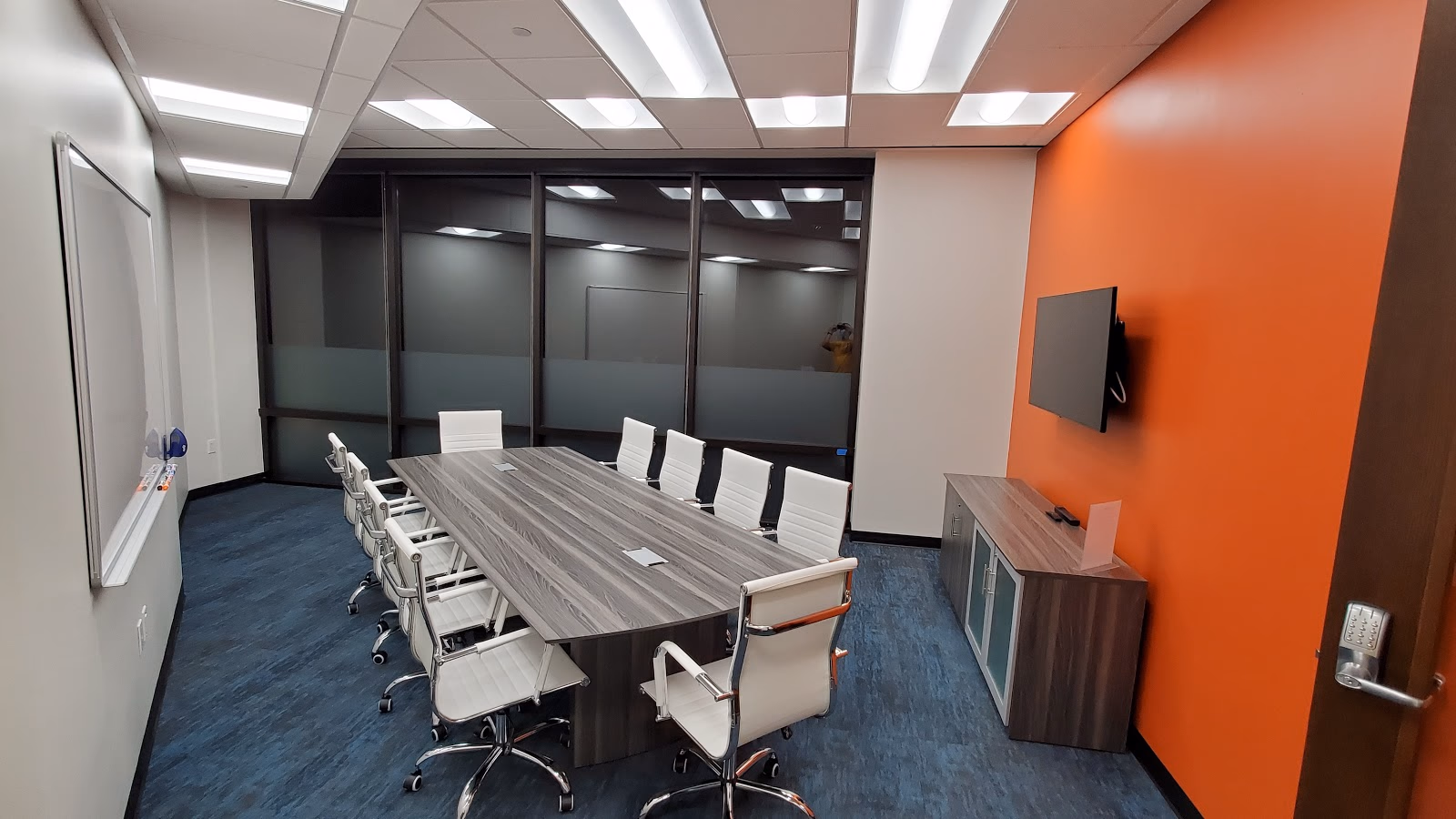 TRUE Space Dallas North corporate conference room with grey wood-grain conference table, white leather executive chairs, bold orange accent wall with wall-mounted TV, frosted glass partition wall, blue commercial carpet, and whiteboard