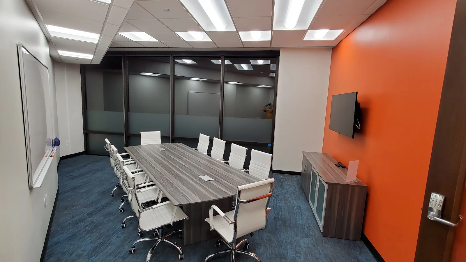 TRUE Space Dallas North corporate conference room with grey wood-grain conference table, white leather executive chairs, bold orange accent wall with wall-mounted TV, frosted glass partition wall, blue commercial carpet, and whiteboard