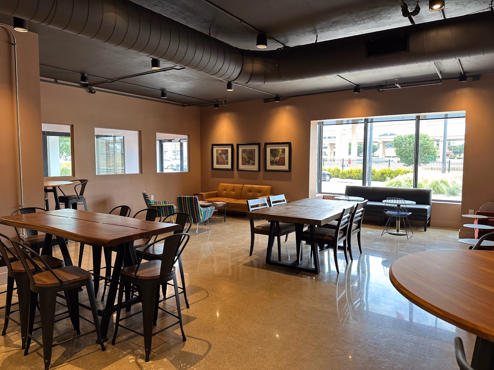 CoWork and Coffee cafe-style interior with warm taupe walls, exposed black ductwork, wood-top high tables with industrial metal chairs, brown leather sofa, striped accent chairs, framed art on walls, large storefront windows, and polished concrete floors