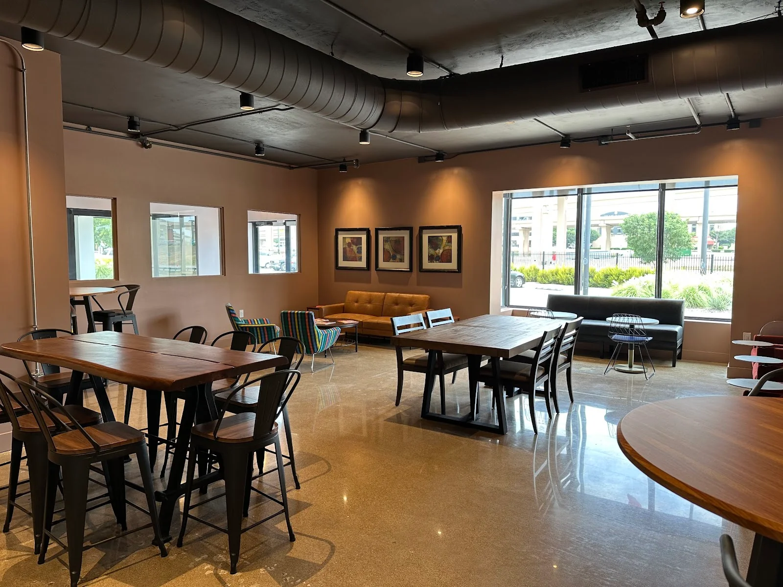 CoWork and Coffee cafe-style interior with warm taupe walls, exposed black ductwork, wood-top high tables with industrial metal chairs, brown leather sofa, striped accent chairs, framed art on walls, large storefront windows, and polished concrete floors